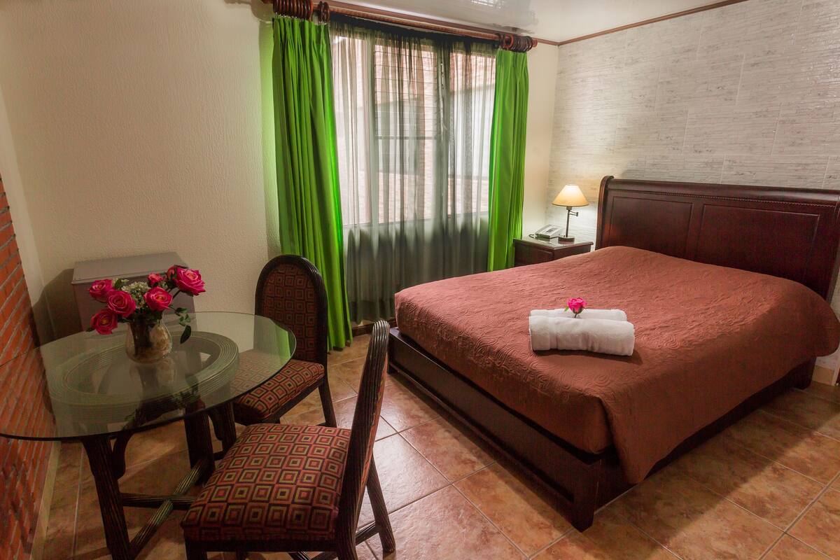 A hotel room with a bed, a round glass table with a vase of flowers, a chair, and a nightstand with a lamp.