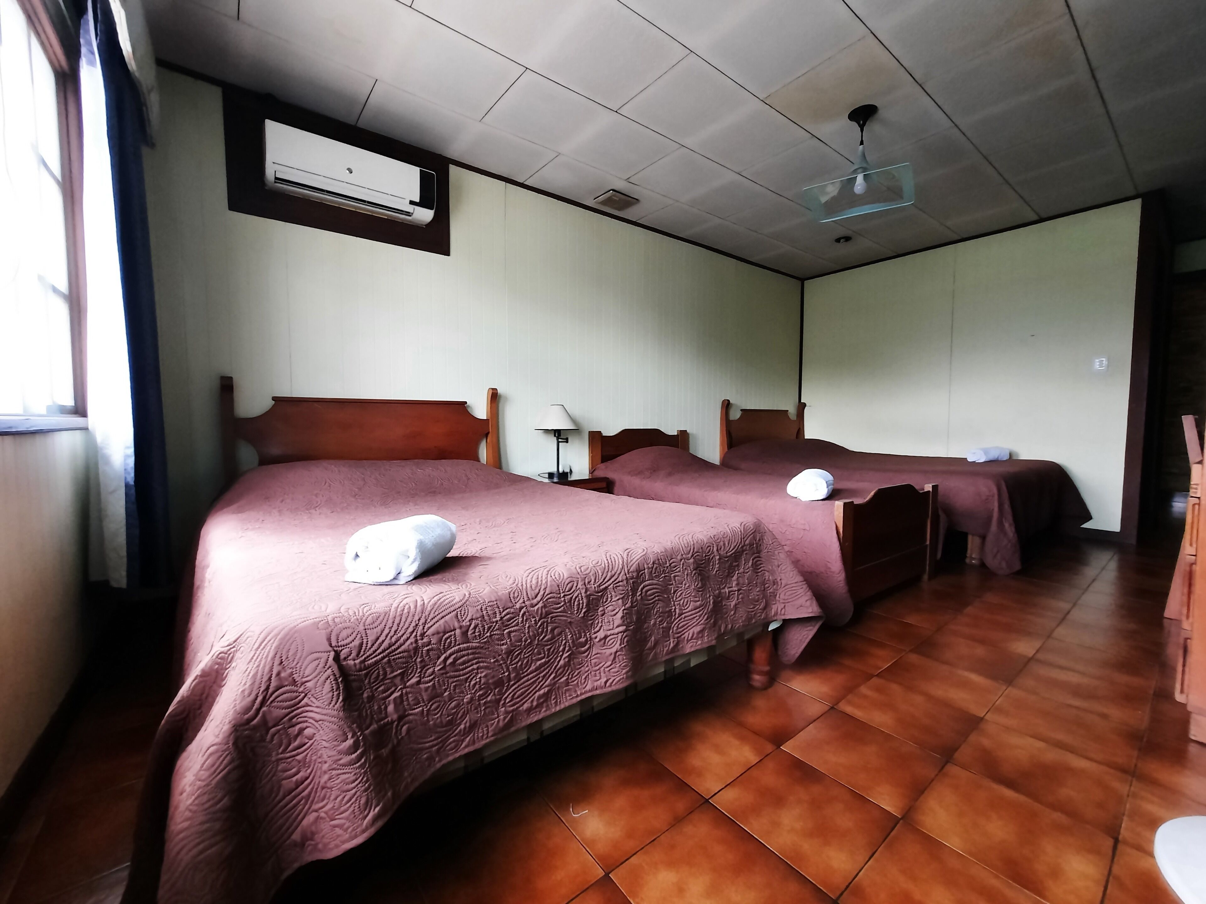 A hotel room with two beds, a ceiling fan, an air conditioning unit, and a window with curtains.