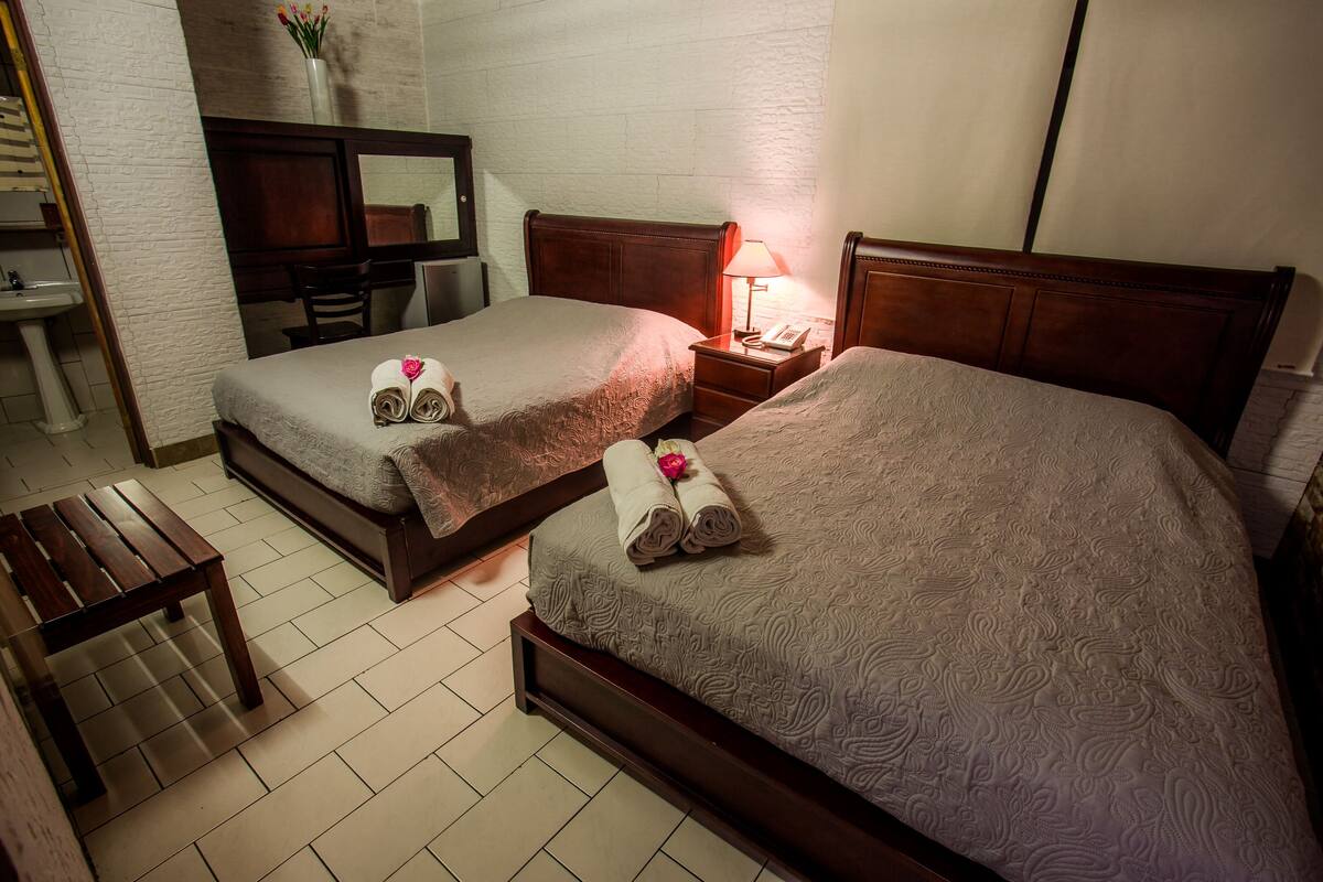 A hotel room with two beds, a wooden bench, a nightstand with a lamp, a mirror, a sink, and a vase with flowers.