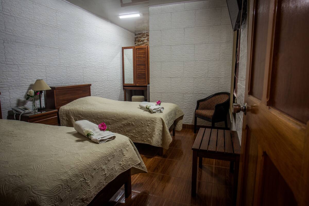 A hotel room with two beds, a wooden chair, a desk with a lamp, and a mirror.