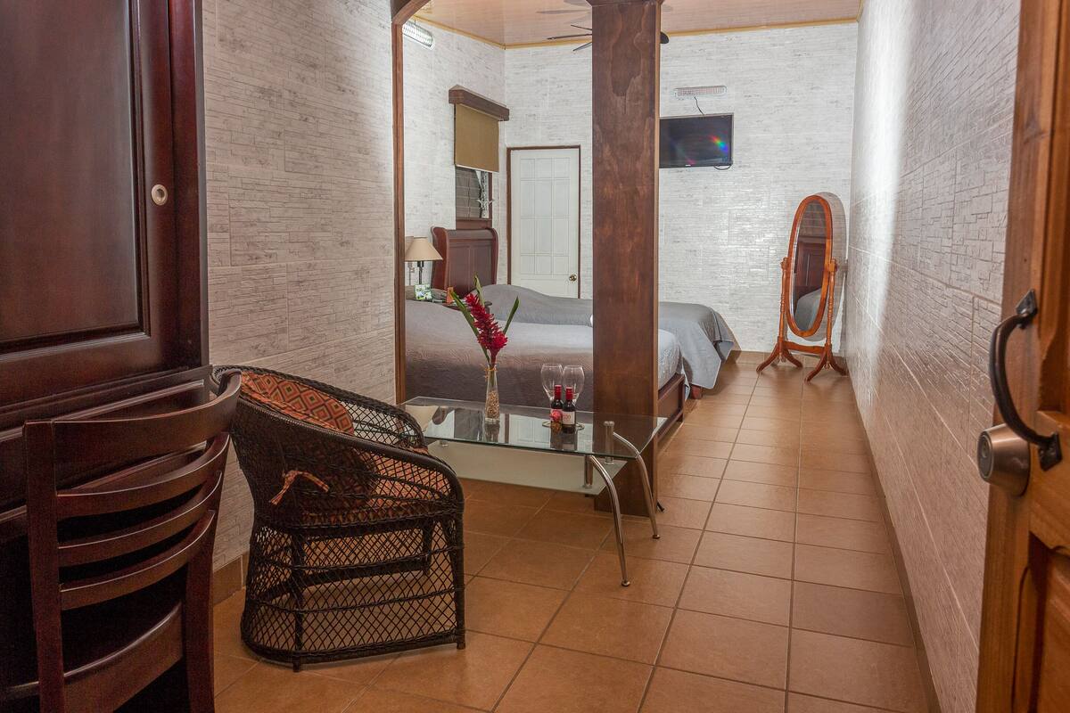 A hotel room with a bed, a glass table with a vase and wine glasses, a wicker chair, a wooden chair, and a mirror on the wall.