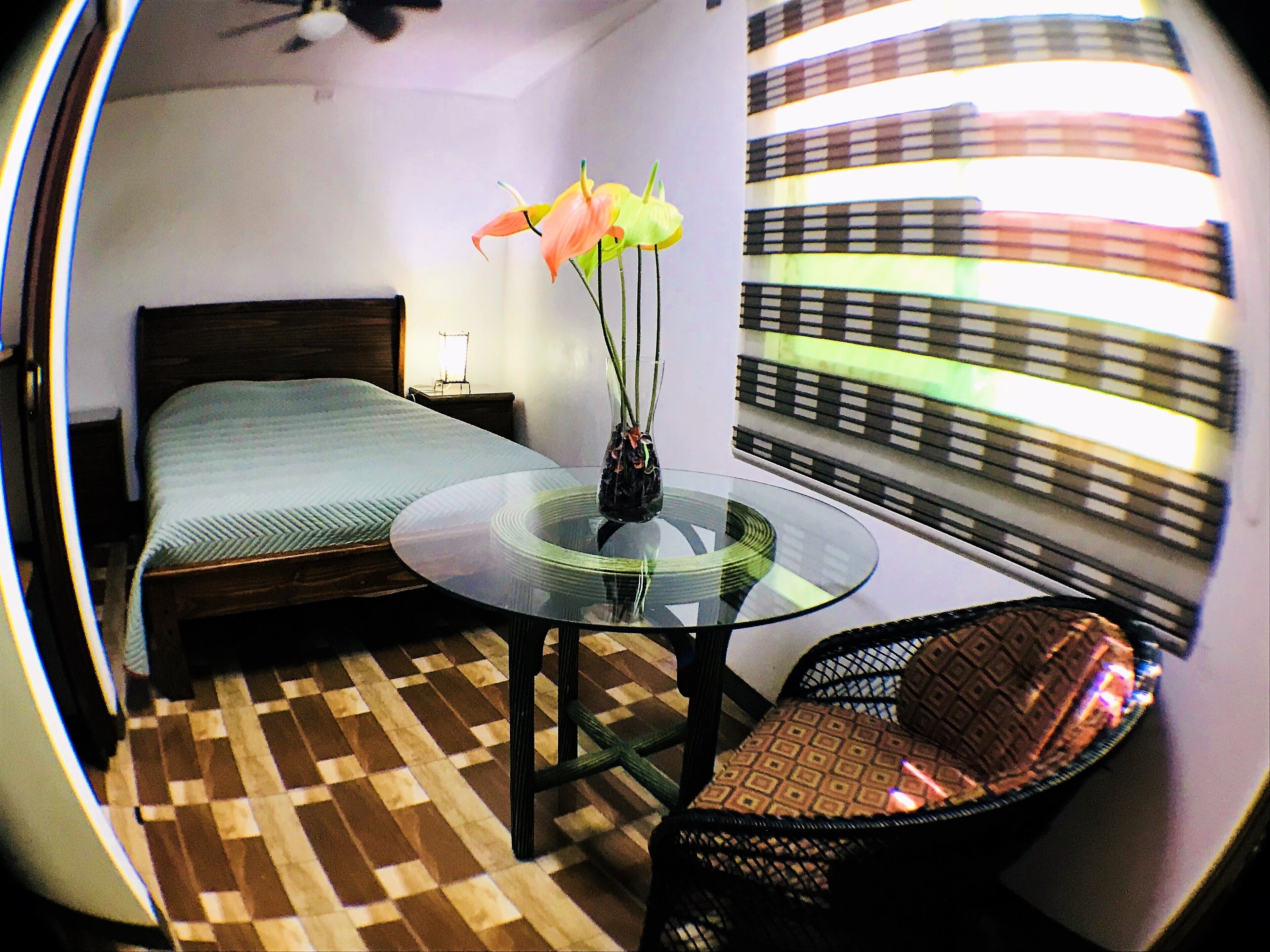 Una habitación de hotel con una cama, una mesa redonda de vidrio con un florero, una silla de mimbre y una ventana con persianas.
