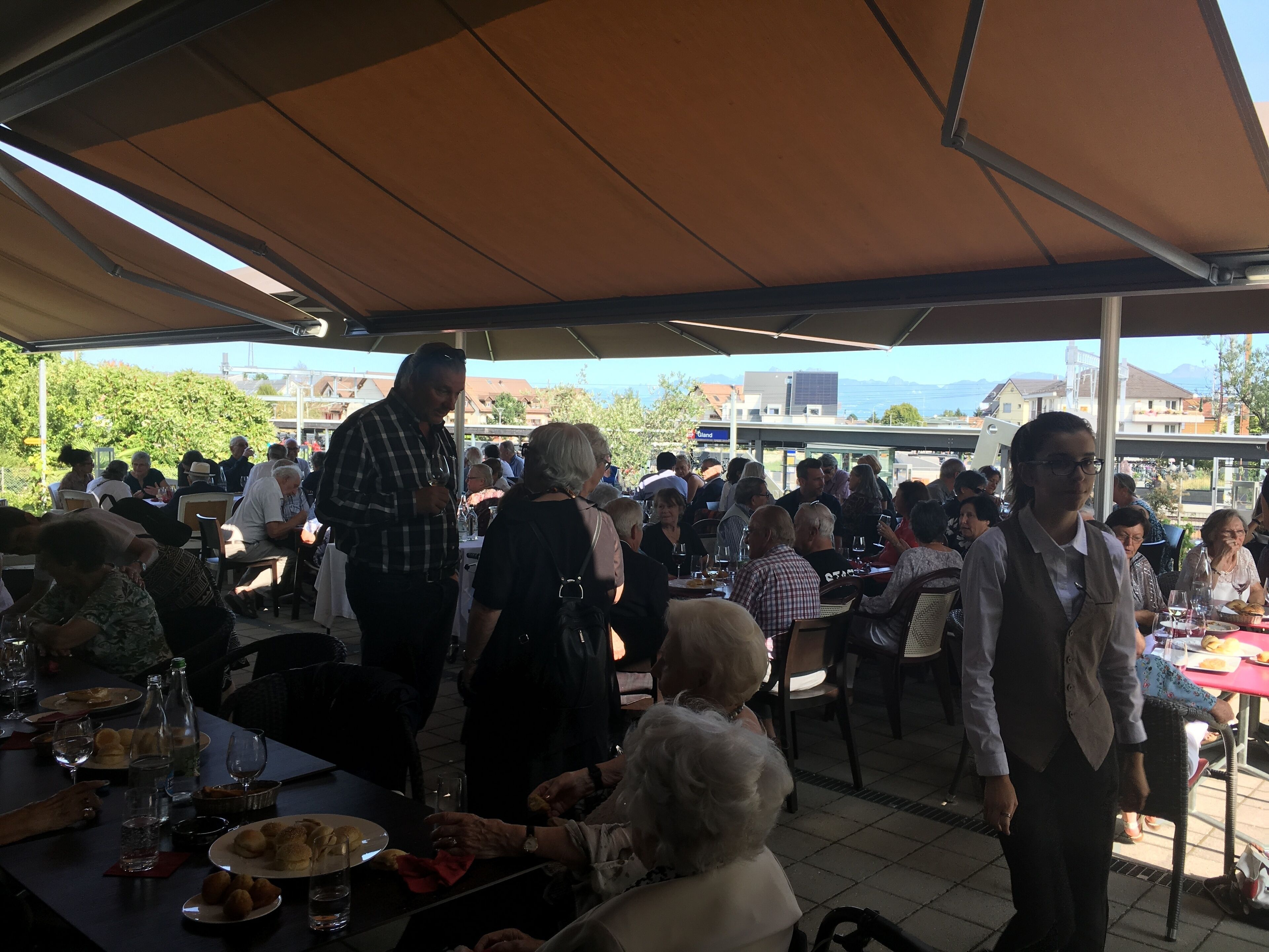 Un área de comedor al aire libre con mesas y sillas, gente sentada y de pie, y vistas de edificios y árboles al fondo.
