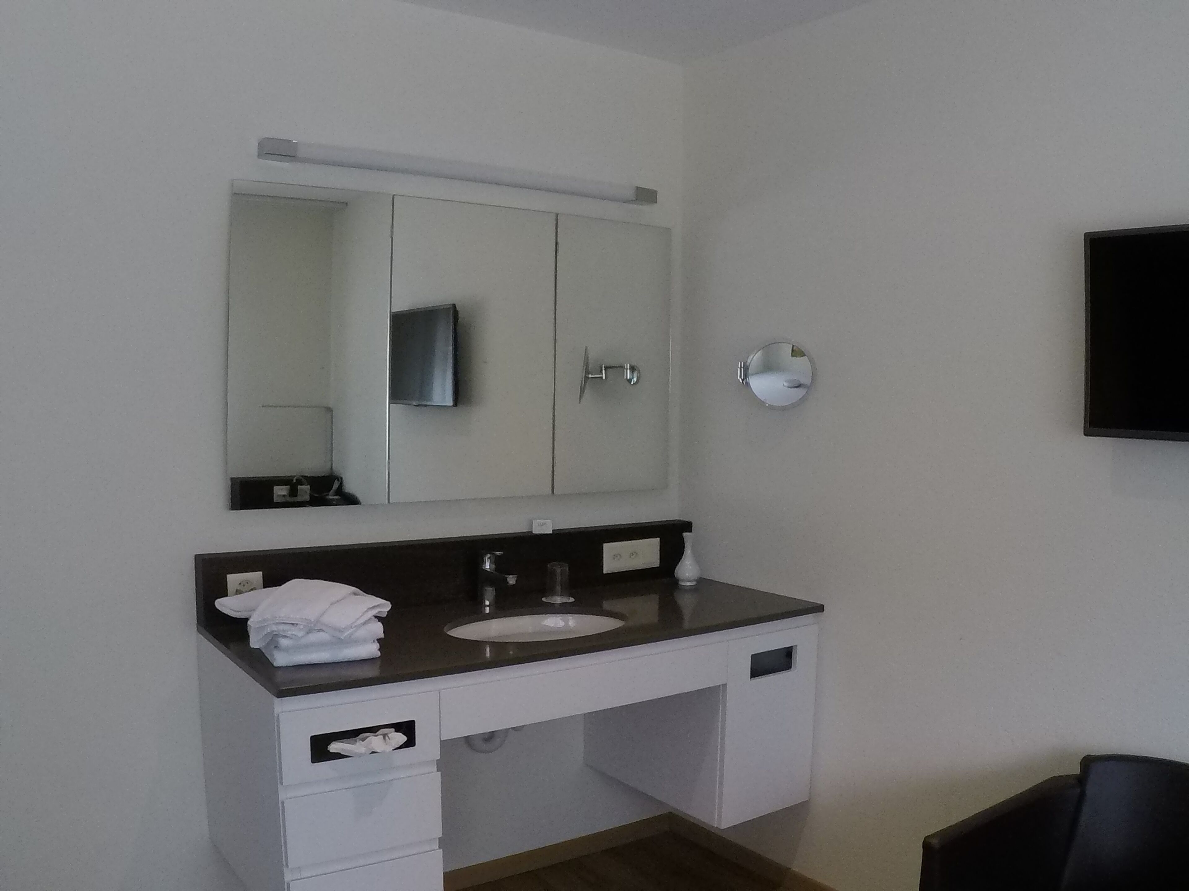 Un baño moderno con un tocador blanco, un espejo grande, un lavamanos y un televisor instalado en la pared.