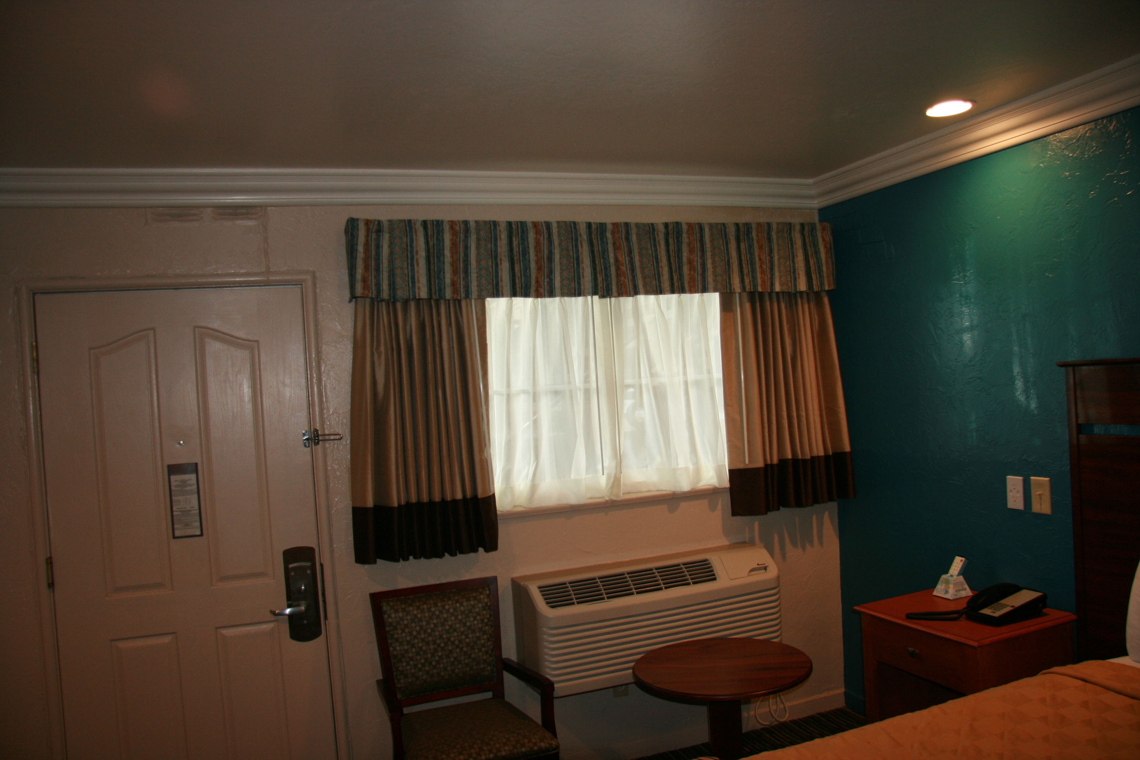 A hotel room with a door, a window with curtains, a chair, a small table, an air conditioning unit, and a bedside table with a lamp and a phone.