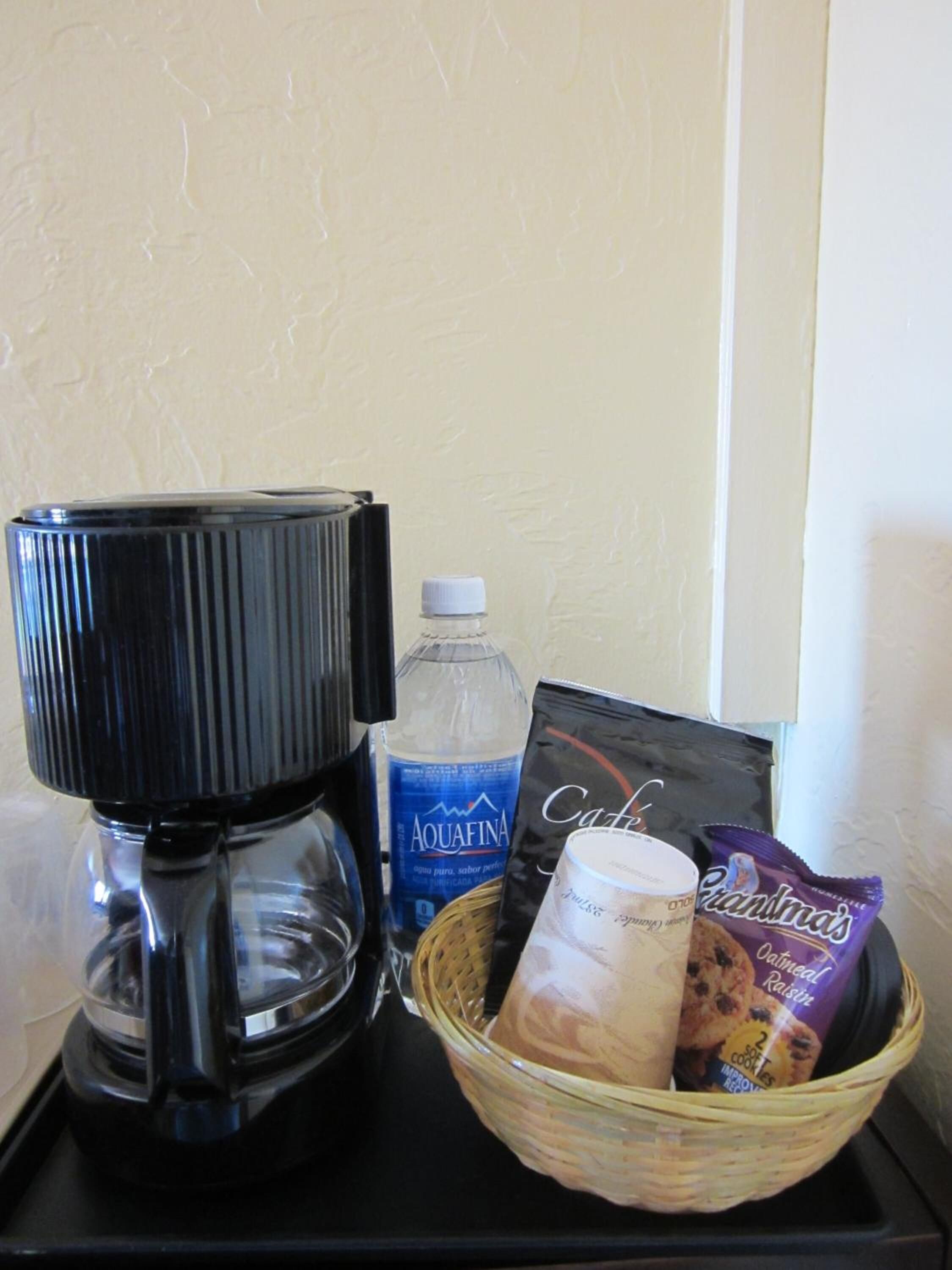A coffee maker, a bottle of Aquafina water, a basket with a candle and snacks, and a card on a black tray.