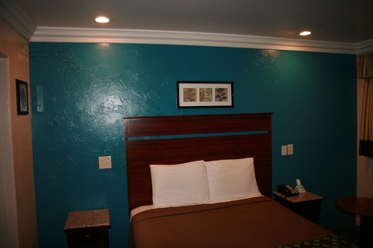 A hotel room with teal walls, a wooden headboard, a bed with white linens, a nightstand with a phone, and a small table with a lamp.