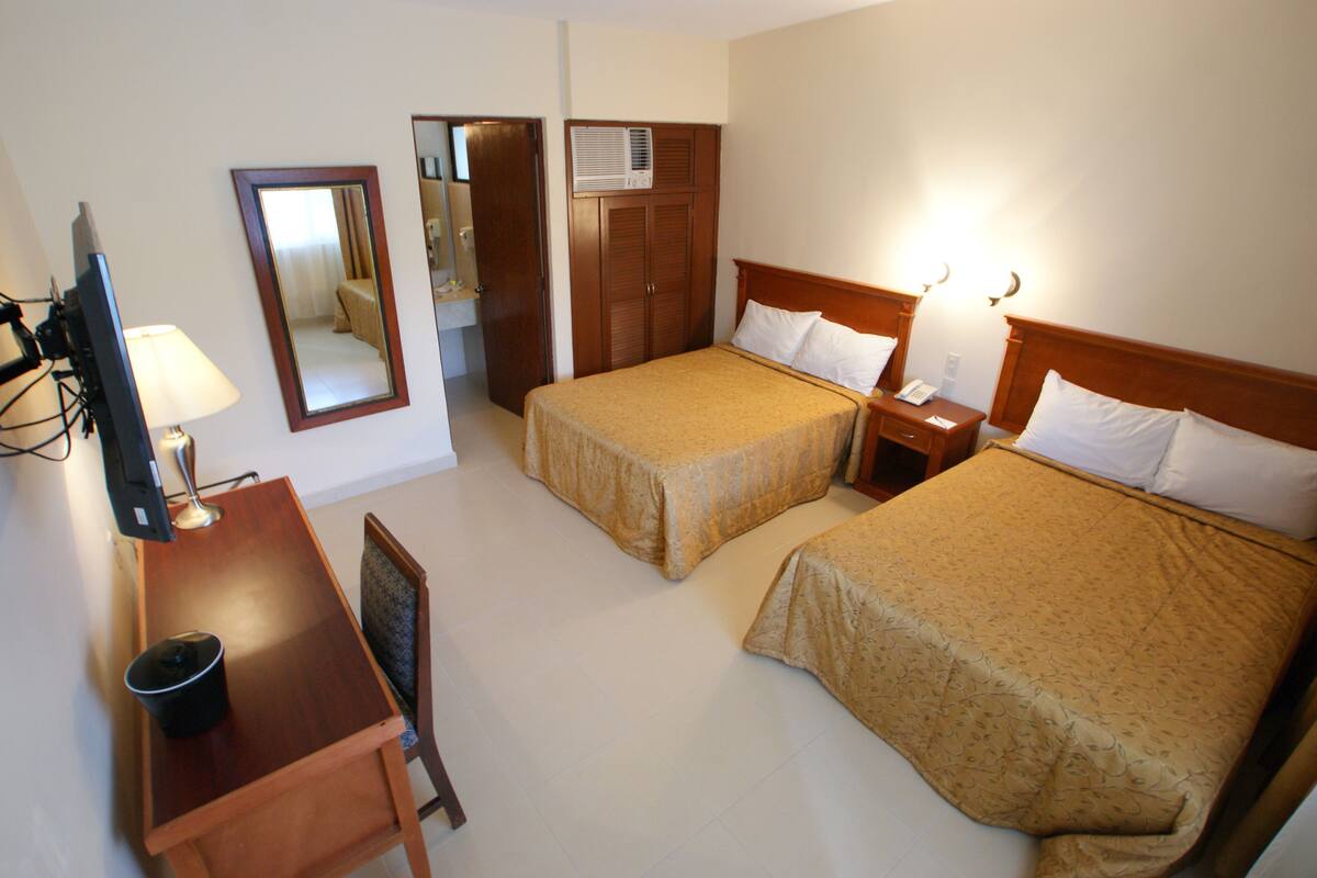 Quarto de hotel com duas camas, uma escrivaninha com luminária, uma cadeira, um espelho e um guarda-roupa.