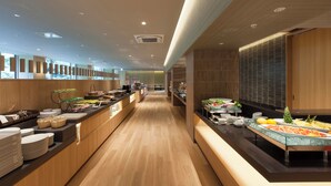 A modern hotel buffet with wooden flooring, a long counter with various food items, and a view of the kitchen in the background. - Hakone Pax Yoshino (Hakone)