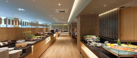 A modern hotel buffet with wooden flooring, a long counter with various food items, and a view of the kitchen in the background.