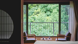 A room with a bed, two chairs, a small table, and a view of greenery through large windows. - Hakone Pax Yoshino (Hakone)