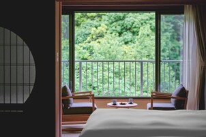 Una habitación con cama, dos sillas, una mesita y vistas a zonas verdes a través de amplios ventanales.