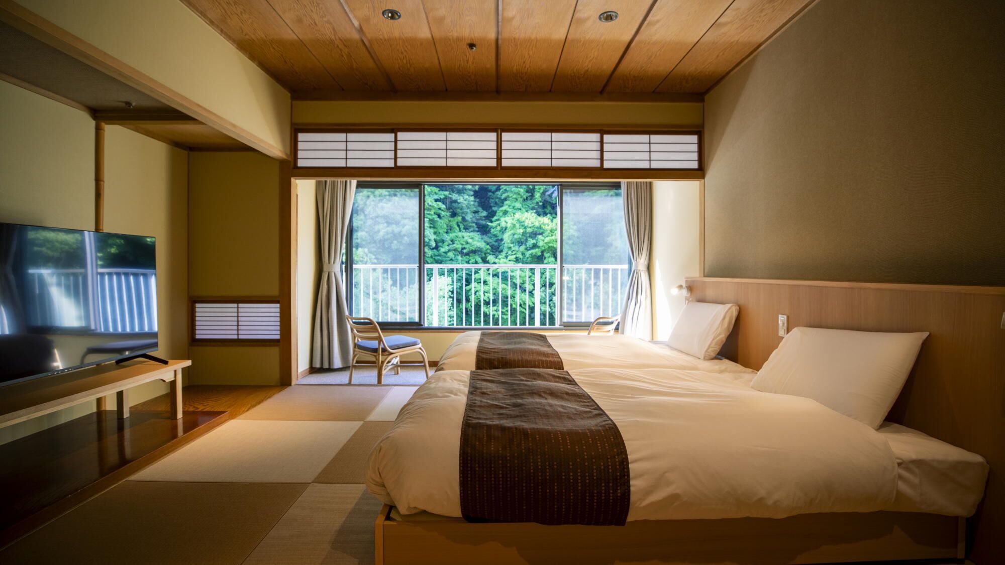 A hotel room with a large bed, a television, a balcony with a view of trees, and sliding doors.