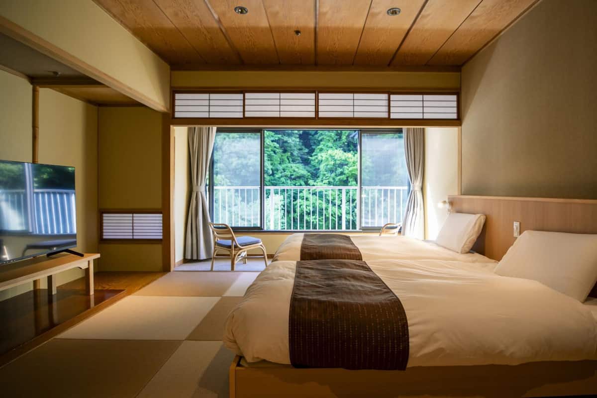 A hotel room with a large bed, a television, a balcony with a view of trees, and sliding doors.
