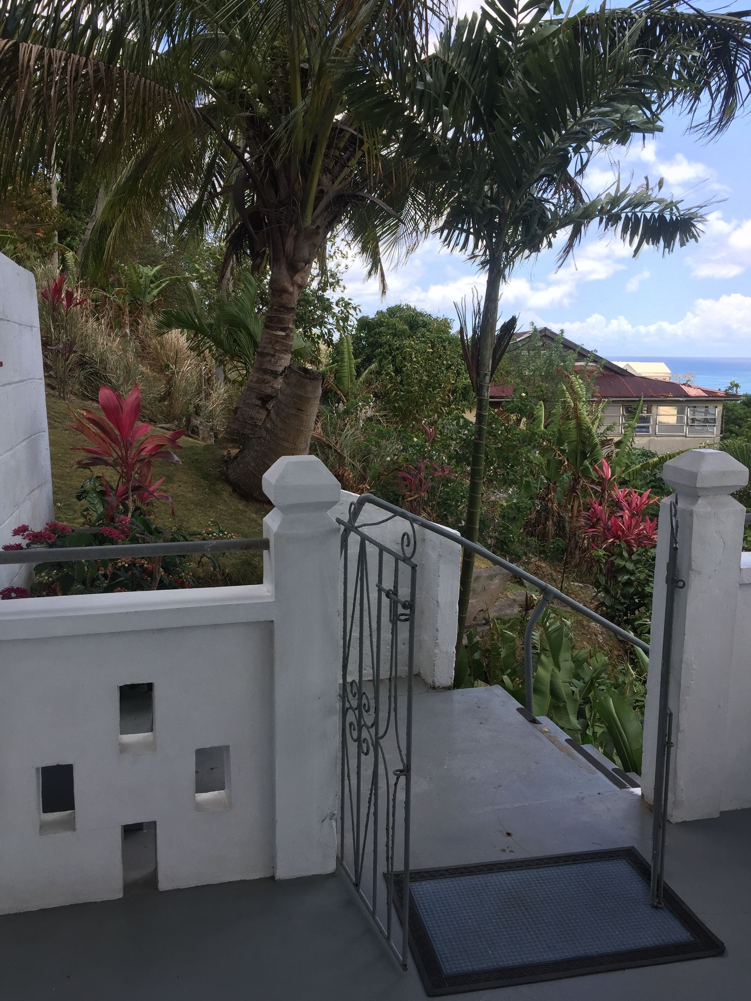 A white staircase leading to a balcony with a metal railing, surrounded by lush greenery and a view of the ocean.