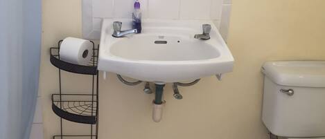 Une salle de bain avec un lavabo blanc, deux robinets, des toilettes et une étagère sur laquelle se trouve un rouleau de papier toilette.