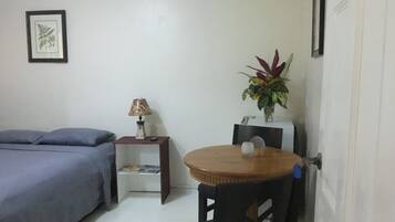 A small, compact room with a bed, a table with a chair, a lamp, and a small plant.