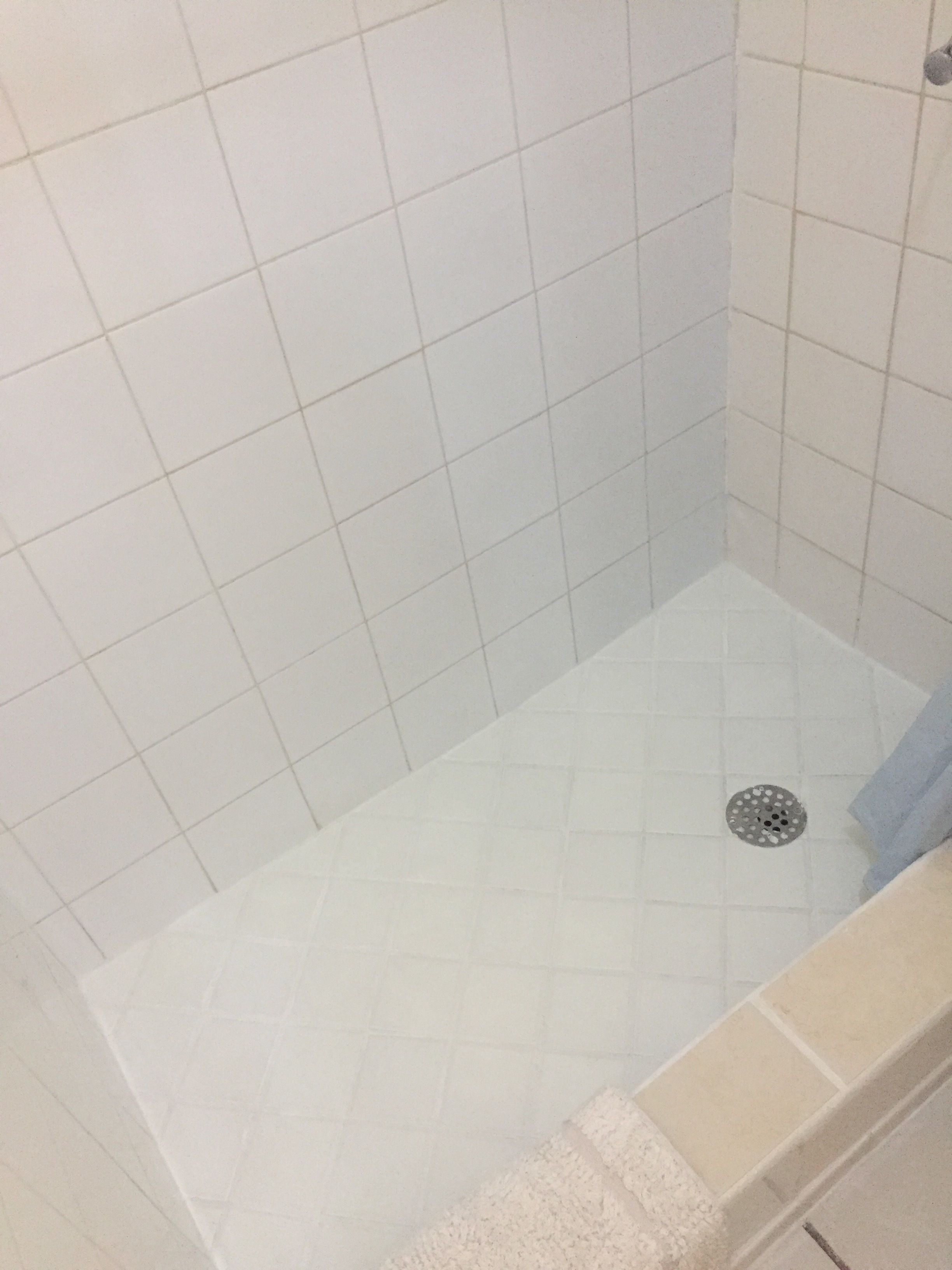 A small, enclosed shower area with white tiled walls and floor, a drain in the center, and a beige bathtub visible to the right.