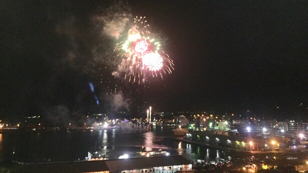 Une vue nocturne d’une ville, illuminée par des feux d’artifice au-dessus d’une étendue d’eau, avec des bâtiments et un pont.
