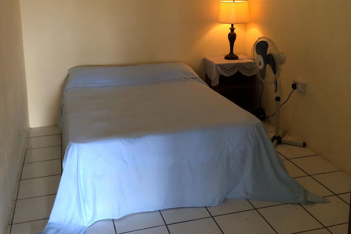 Une petite chambre avec un lit simple, recouverte d’une courtepointe blanche, une table de chevet avec une lampe et un tableau accroché au mur.