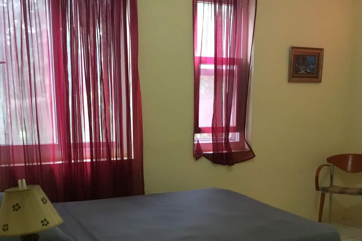 Une pièce comprenant un lit, une chaise, une table avec une lampe et une fenêtre avec des rideaux.