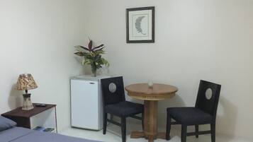 Ein Schlafzimmer mit einem Bett, einem kleinen Tisch mit Lampe, einem Kühlschrank, zwei Stühlen, einem runden Tisch und einem gerahmten Bild an der Wand.