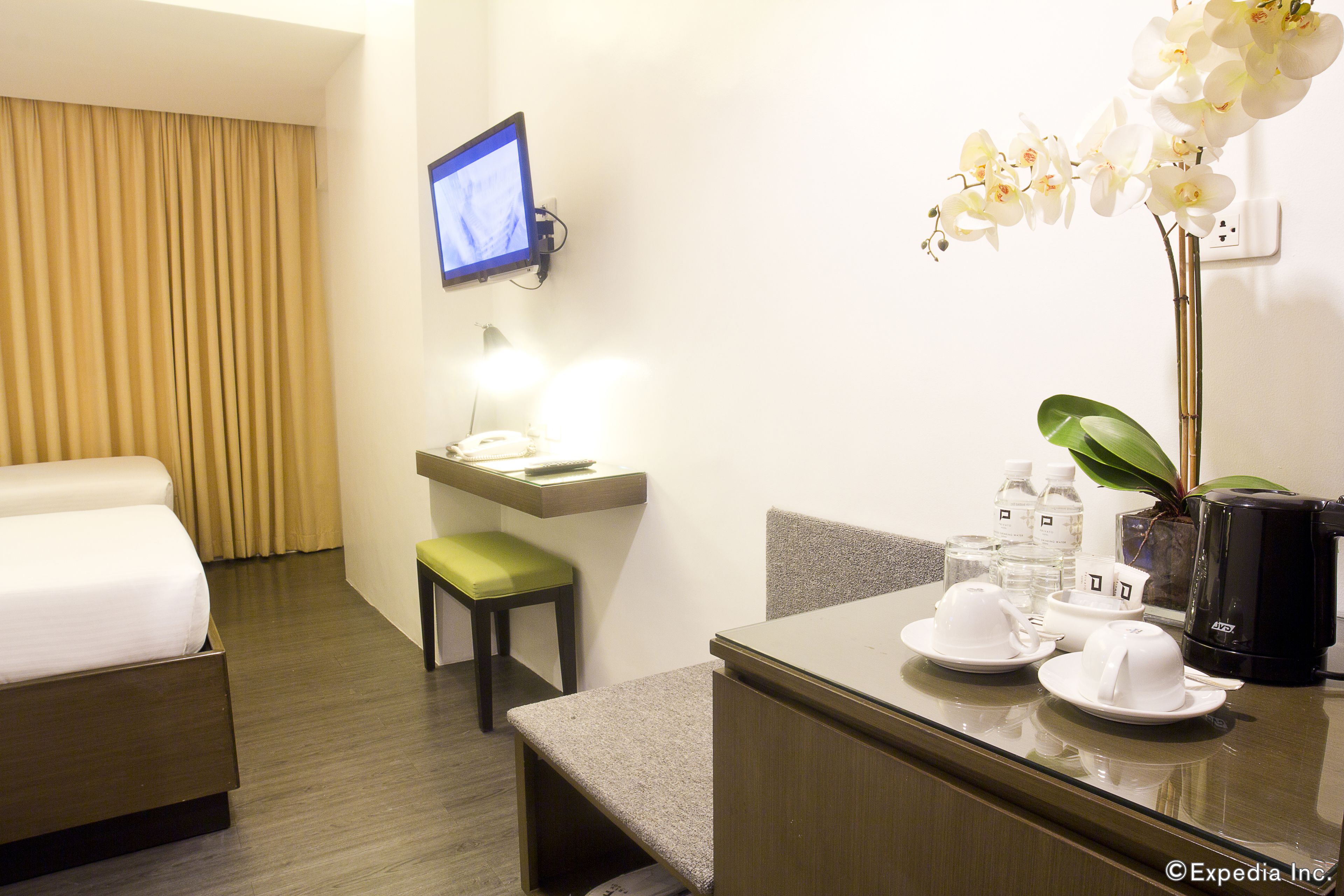 Habitación de hotel con cama, escritorio con monitor de ordenador, silla, mesita con cafetera y una orquídea en maceta.