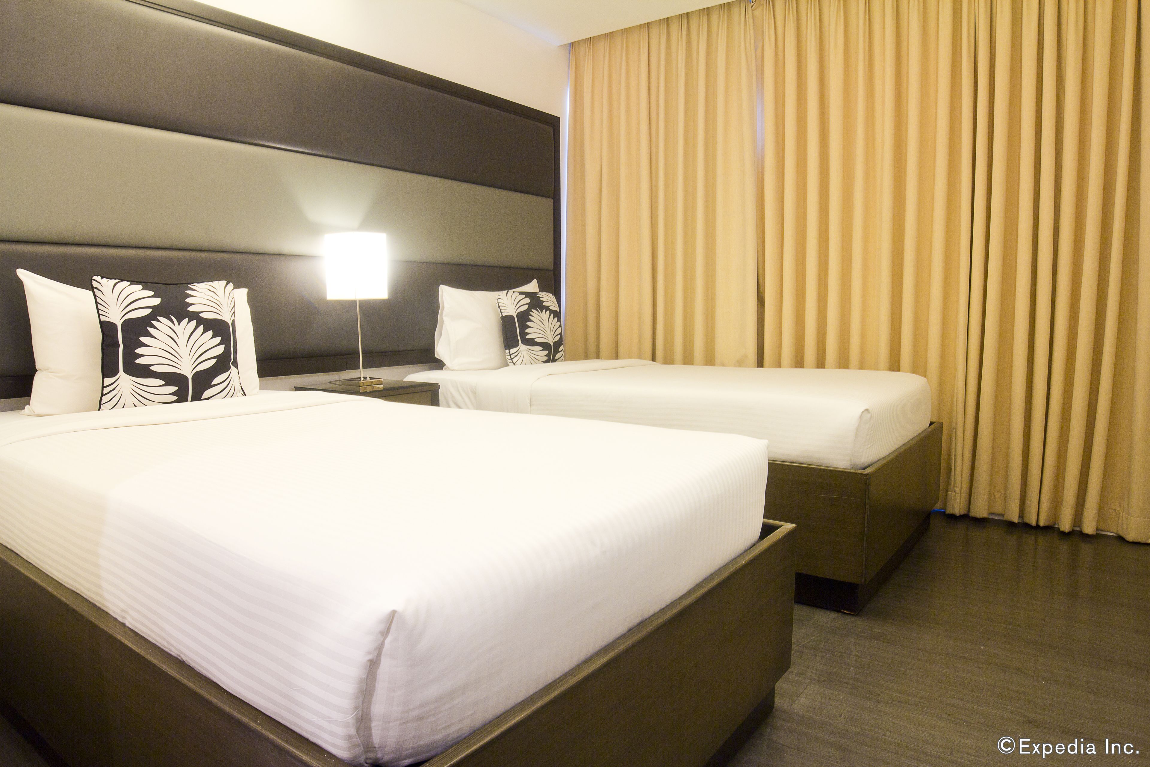Ein Hotelzimmer mit zwei Betten, einem Kopfteil, einem Nachttisch, einer Lampe und beige farbenen Vorhängen.