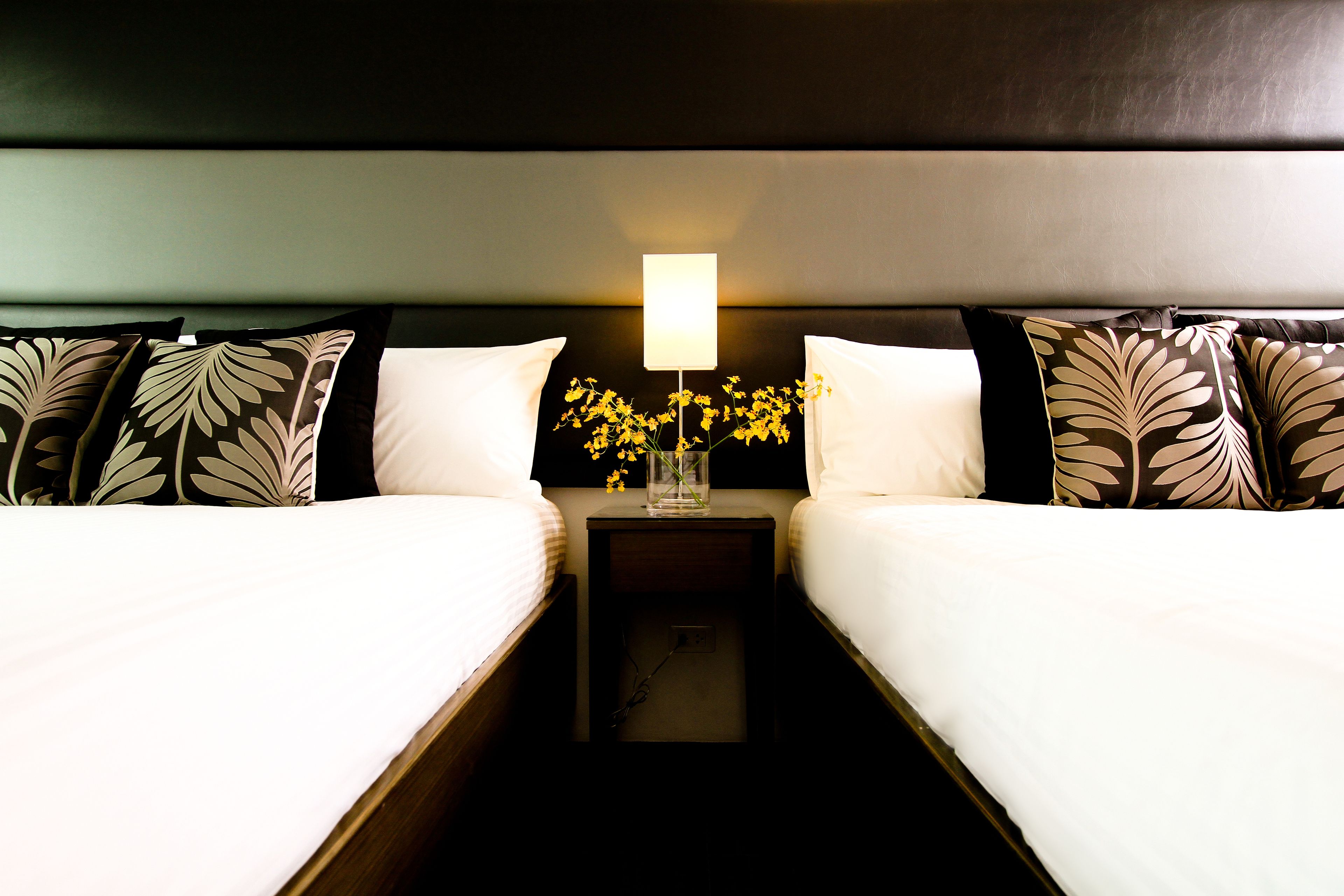 Quarto de hotel com duas camas, uma mesinha de cabeceira com abajur e um vaso com flores.
