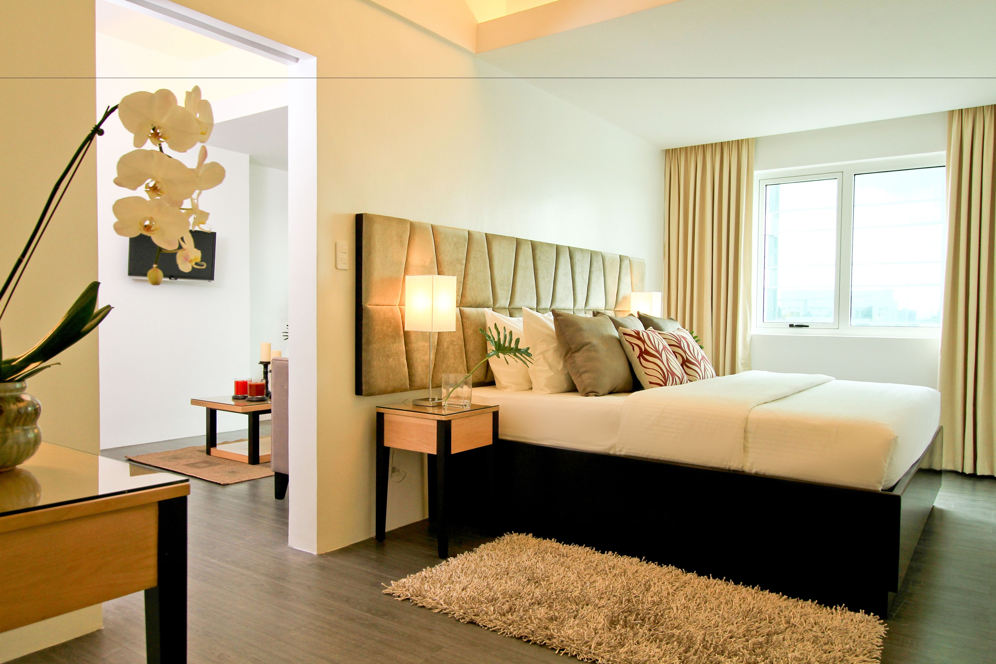 A modern hotel room with a large bed, bedside lamps, a small table with a vase, and a view of the city through the window.