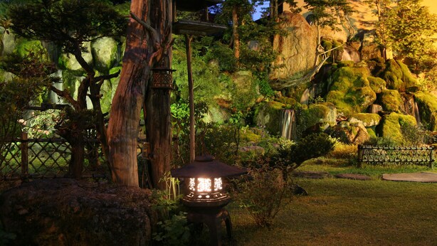 A serene garden with a traditional Japanese lantern, a wooden fence, and a waterfall. - Ryokan Sensyoen (Asahikawa)