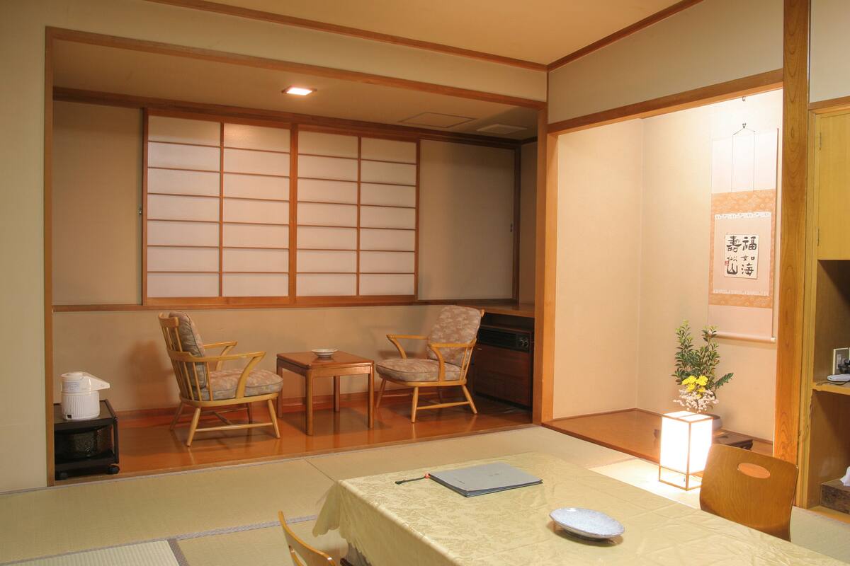 Une pièce de style japonais traditionnel, avec un sol recouvert de tatamis, une petite table basse avec un coussin, deux chaises en bois, une petite table sur laquelle se trouve un bol, et un vase rempli de fleurs.