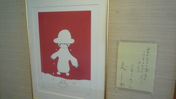 Uma obra de arte emoldurada com fundo vermelho e uma figura branca, possivelmente uma boneca, com chapéu e um objeto pequeno abaixo.