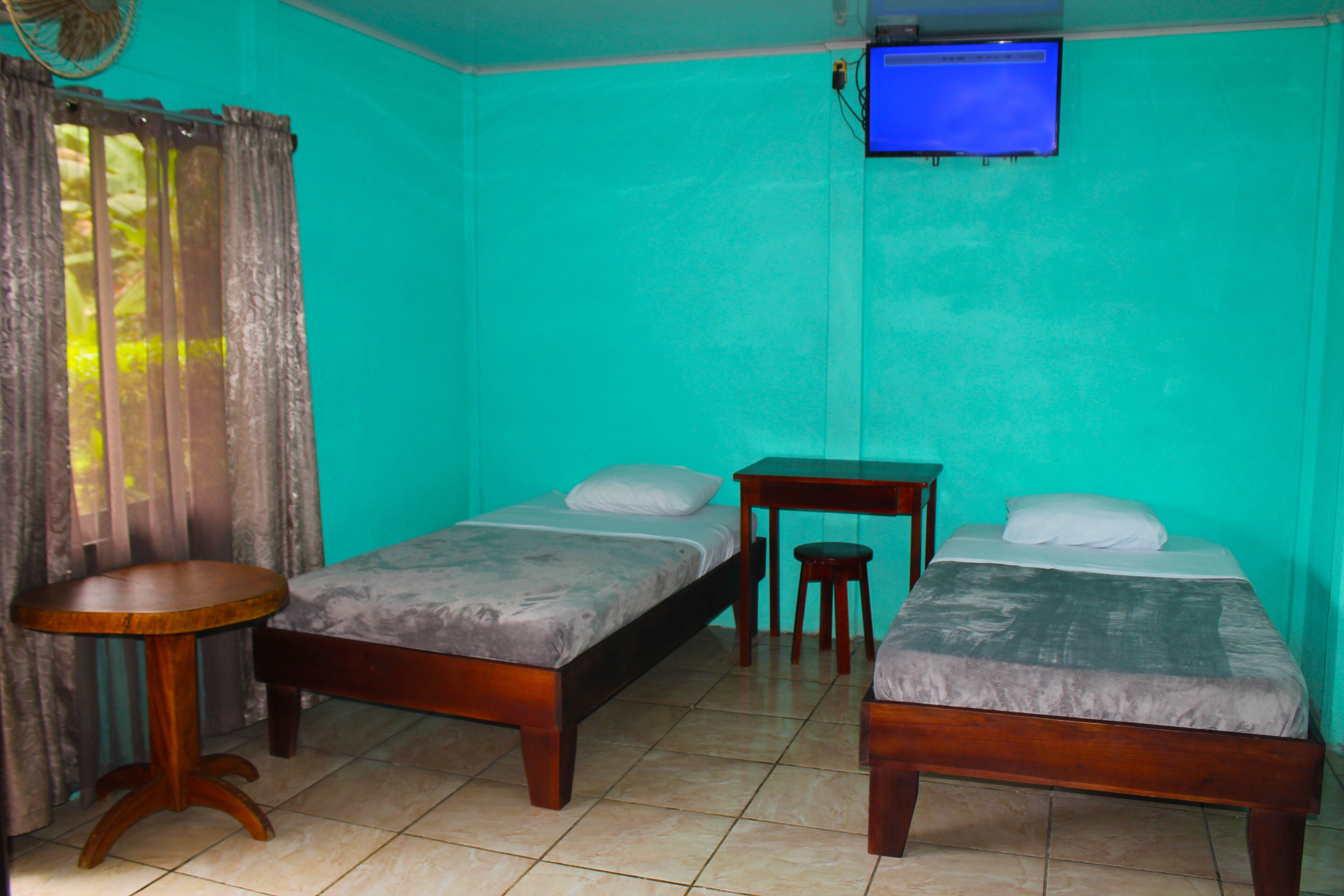 A room with two single beds, a small wooden table, a TV mounted on the wall, and a curtain partially drawn to the side.