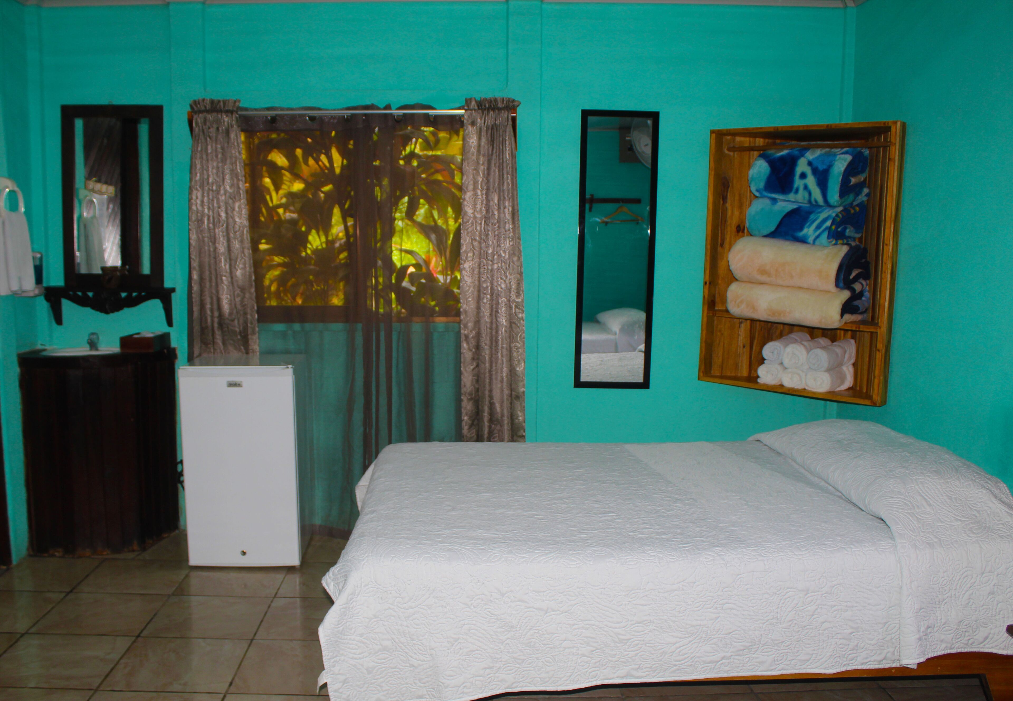 A room with a bed, a small refrigerator, a mirror, and a window with curtains.