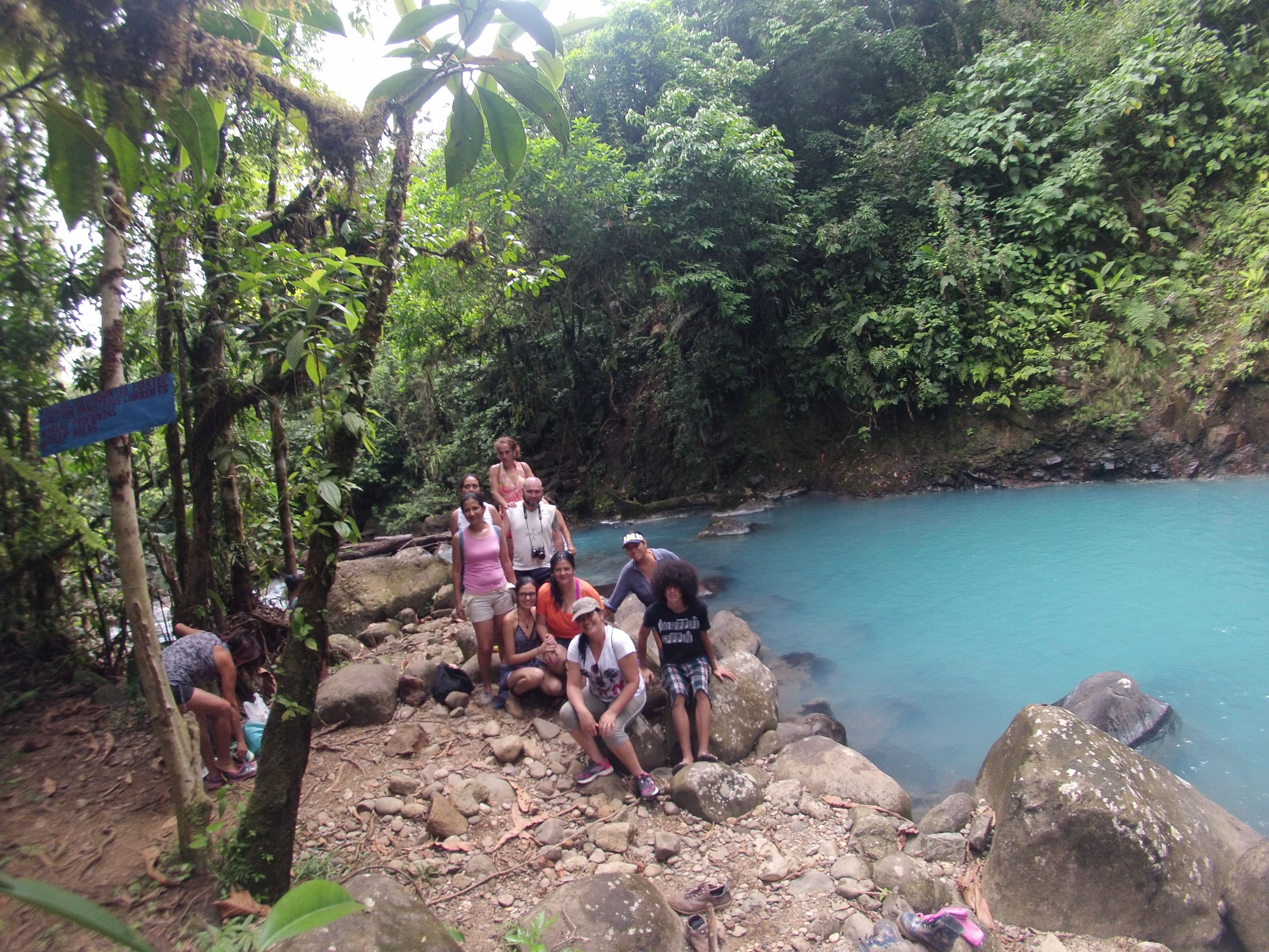 A group of people sitting on rocks by a clear blue water body surrounded by dense greenery.