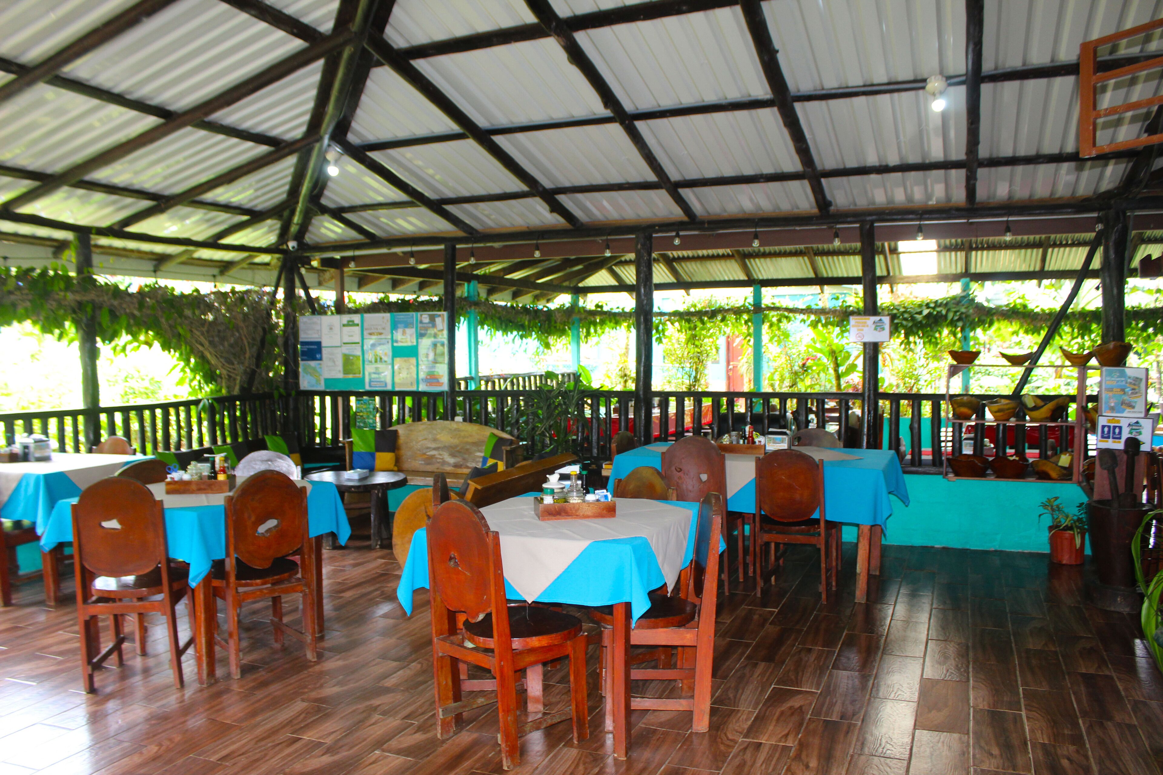 An indoor restaurant with wooden tables and chairs, covered with blue tablecloths, and a tiled floor.