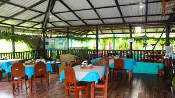 An indoor restaurant with wooden tables and chairs, covered with blue tablecloths, and a tiled floor.
