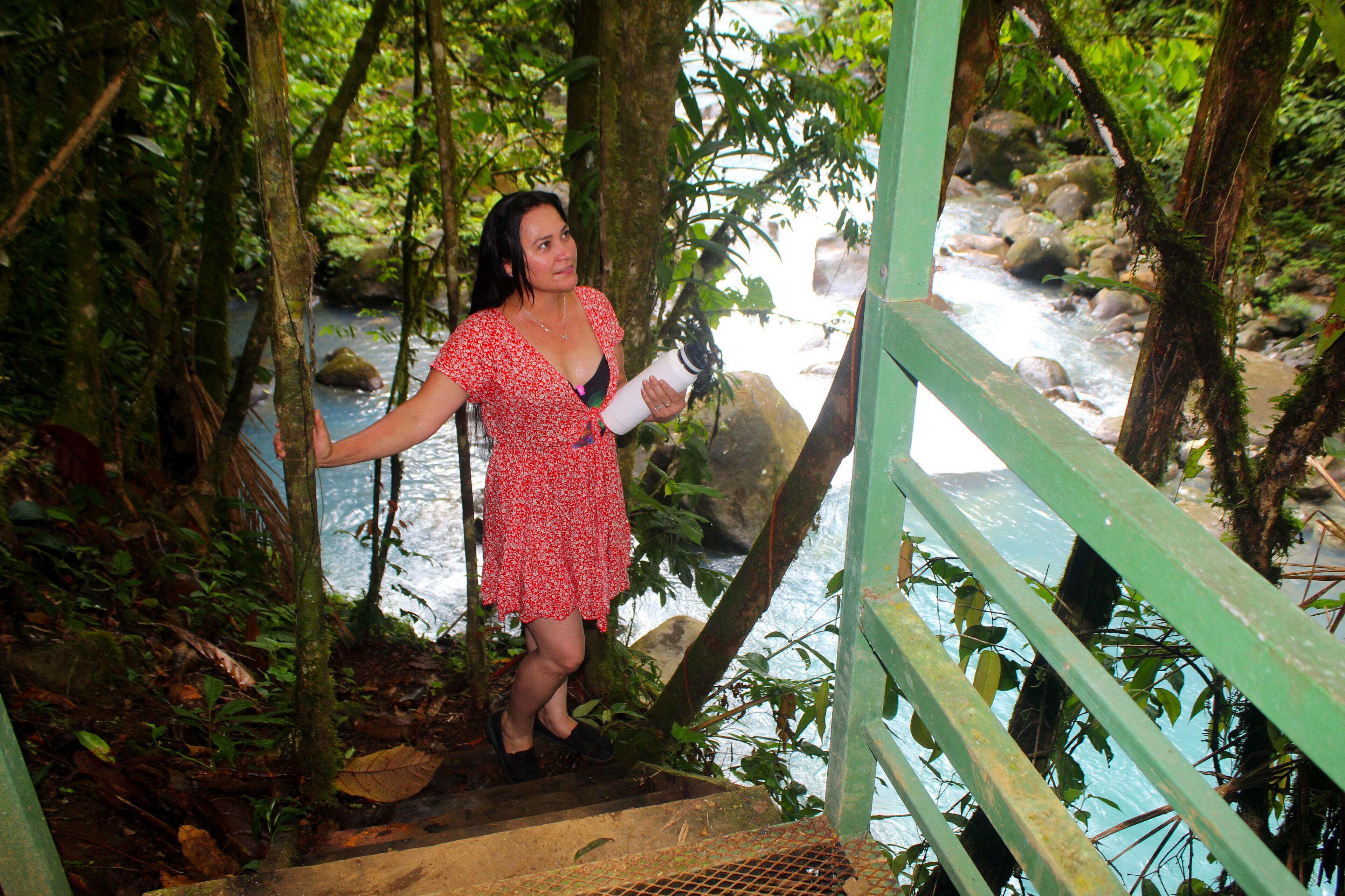 Una mujer con un vestido rojo parada sobre un puente de madera sobre un río rodeado de vegetación densa.