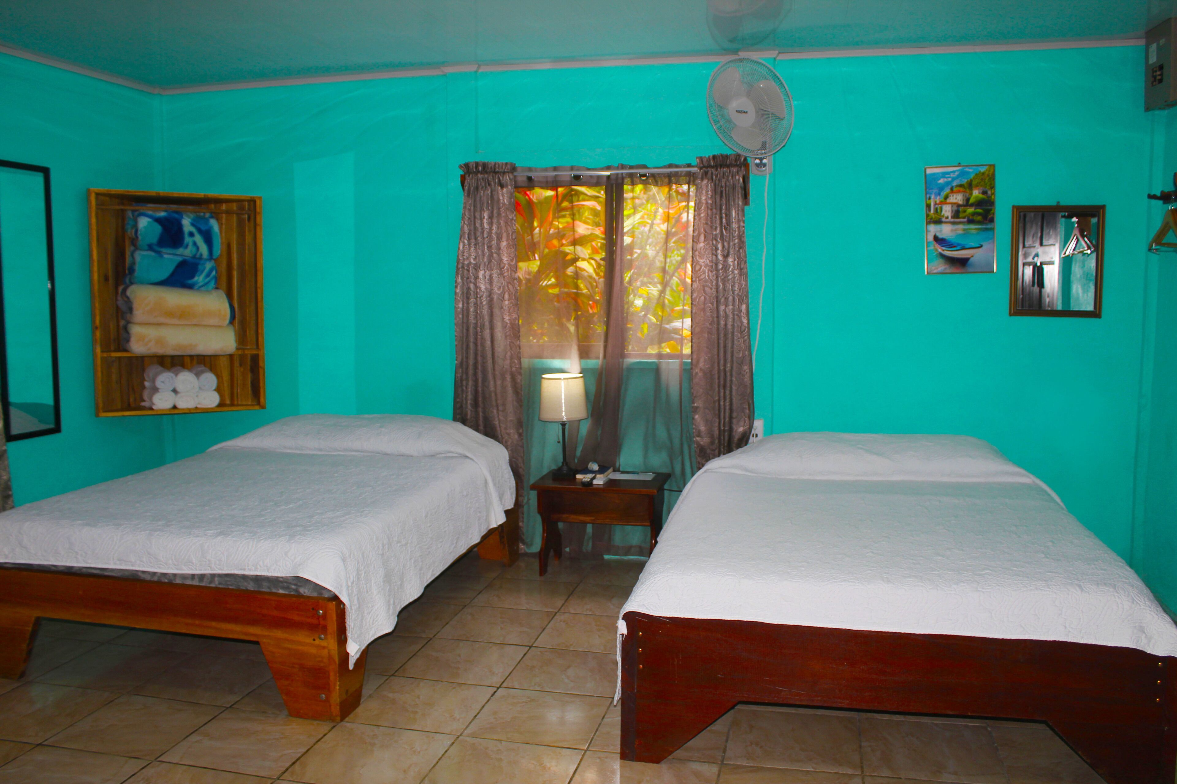 A room with two wooden beds, a fan, a window with curtains, a shelf with towels, and a painting on the wall.