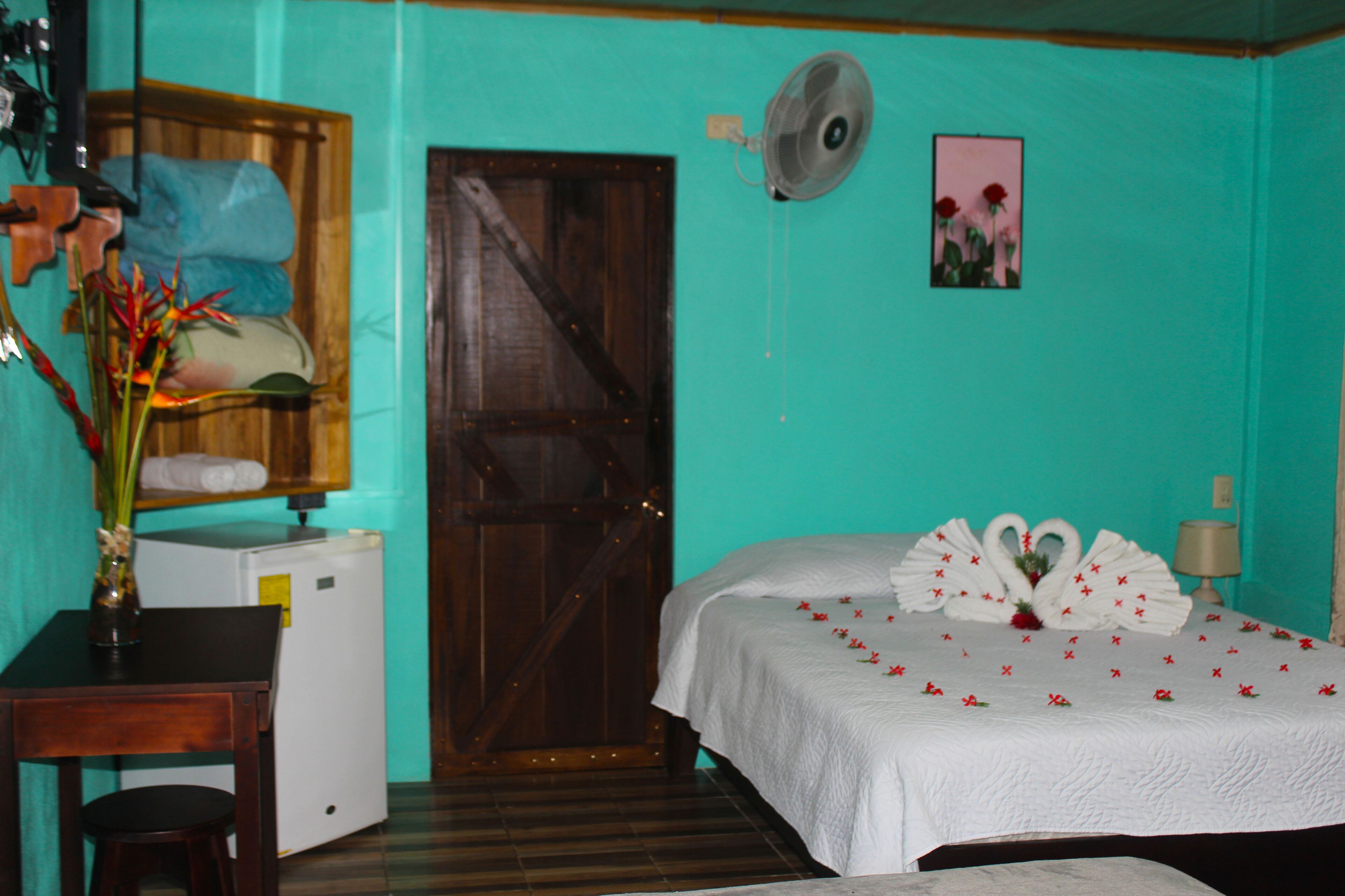 Una habitación con cama, escritorio, refrigerador pequeño, puerta de madera, ventilador de pared y un cuadro de rosas.