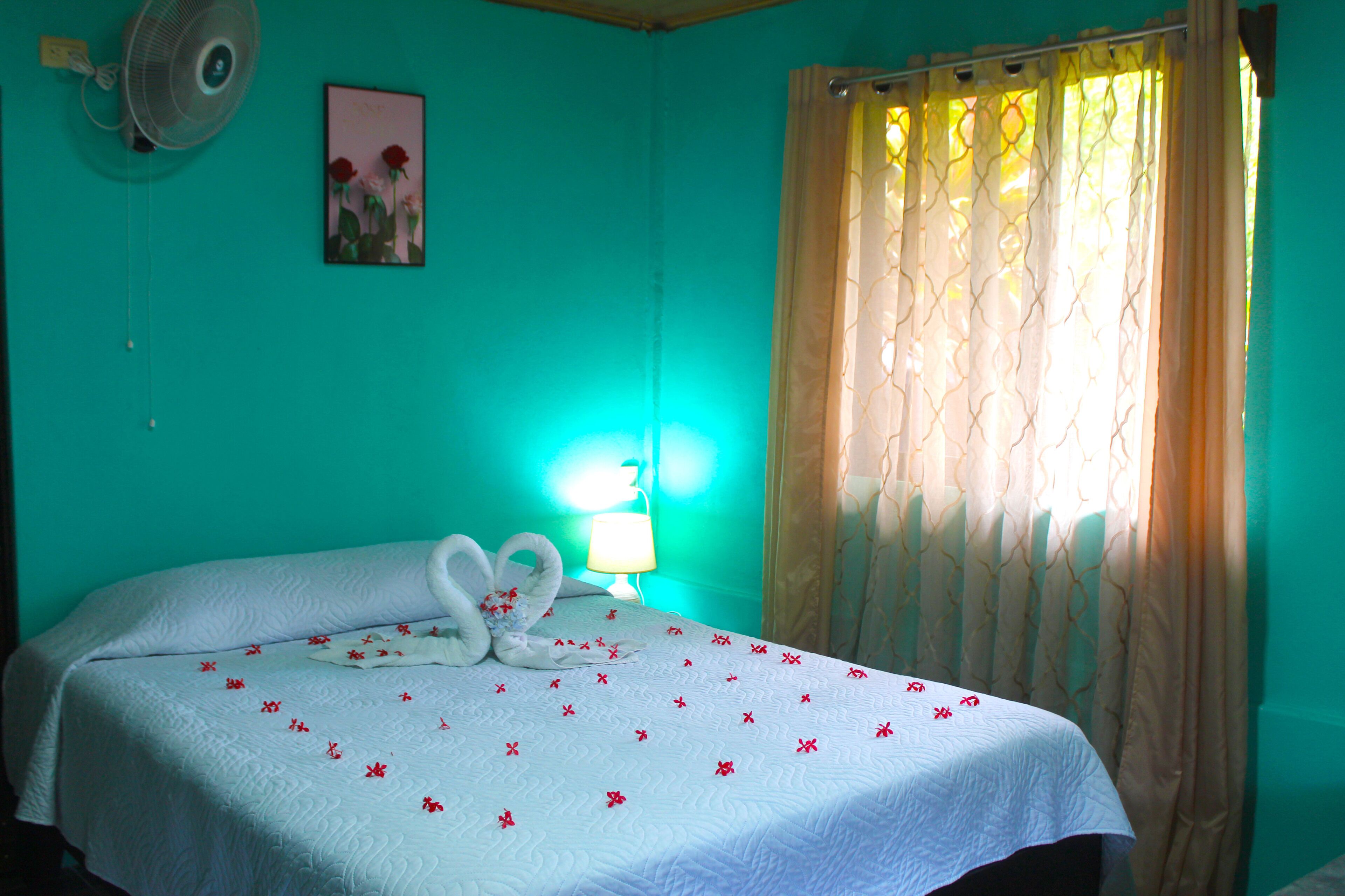 A room with a bed, a wall-mounted fan, a picture of flowers, and a window with curtains.