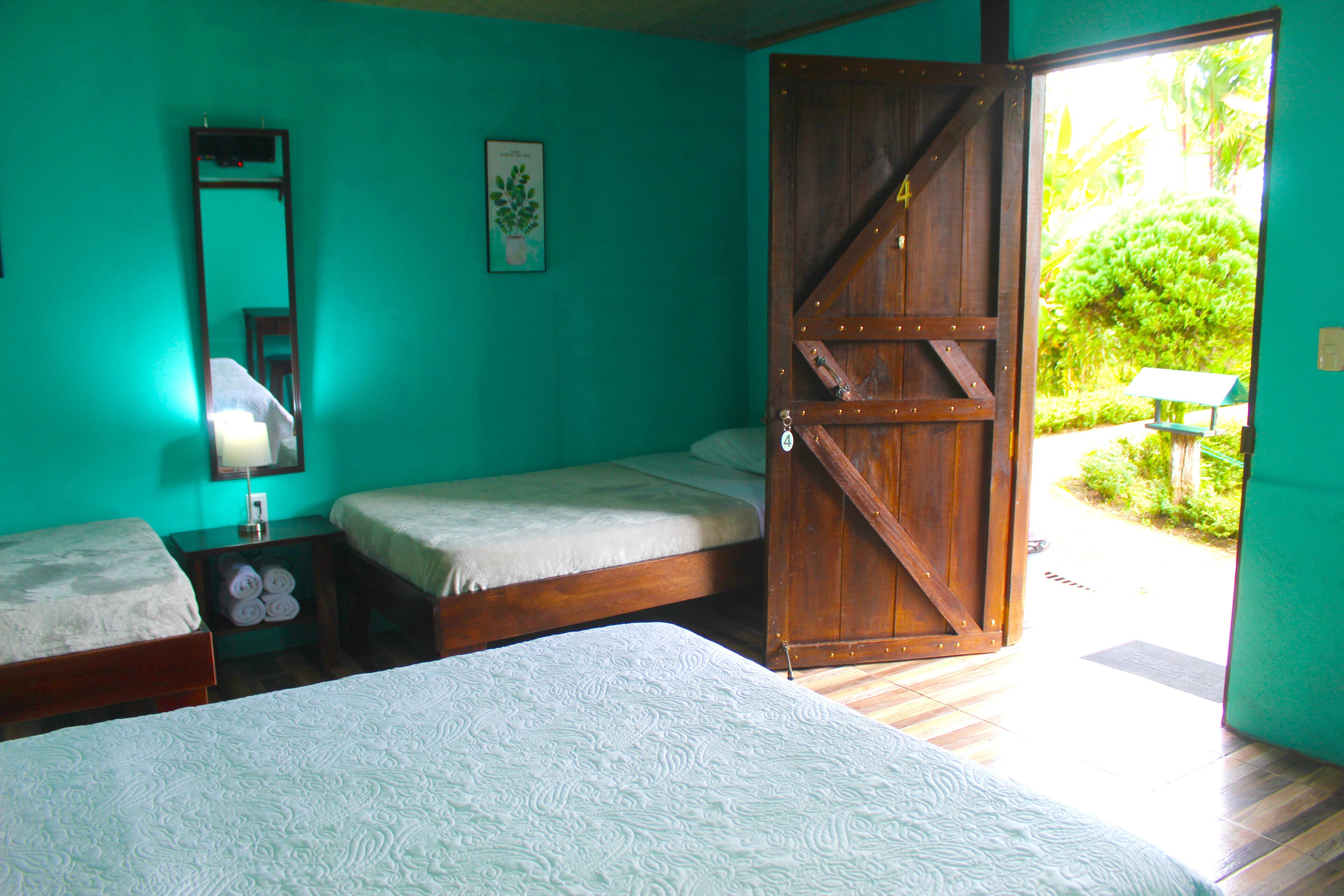 A room with two beds, a wooden door, a small table with a lamp, and a framed picture on the wall.