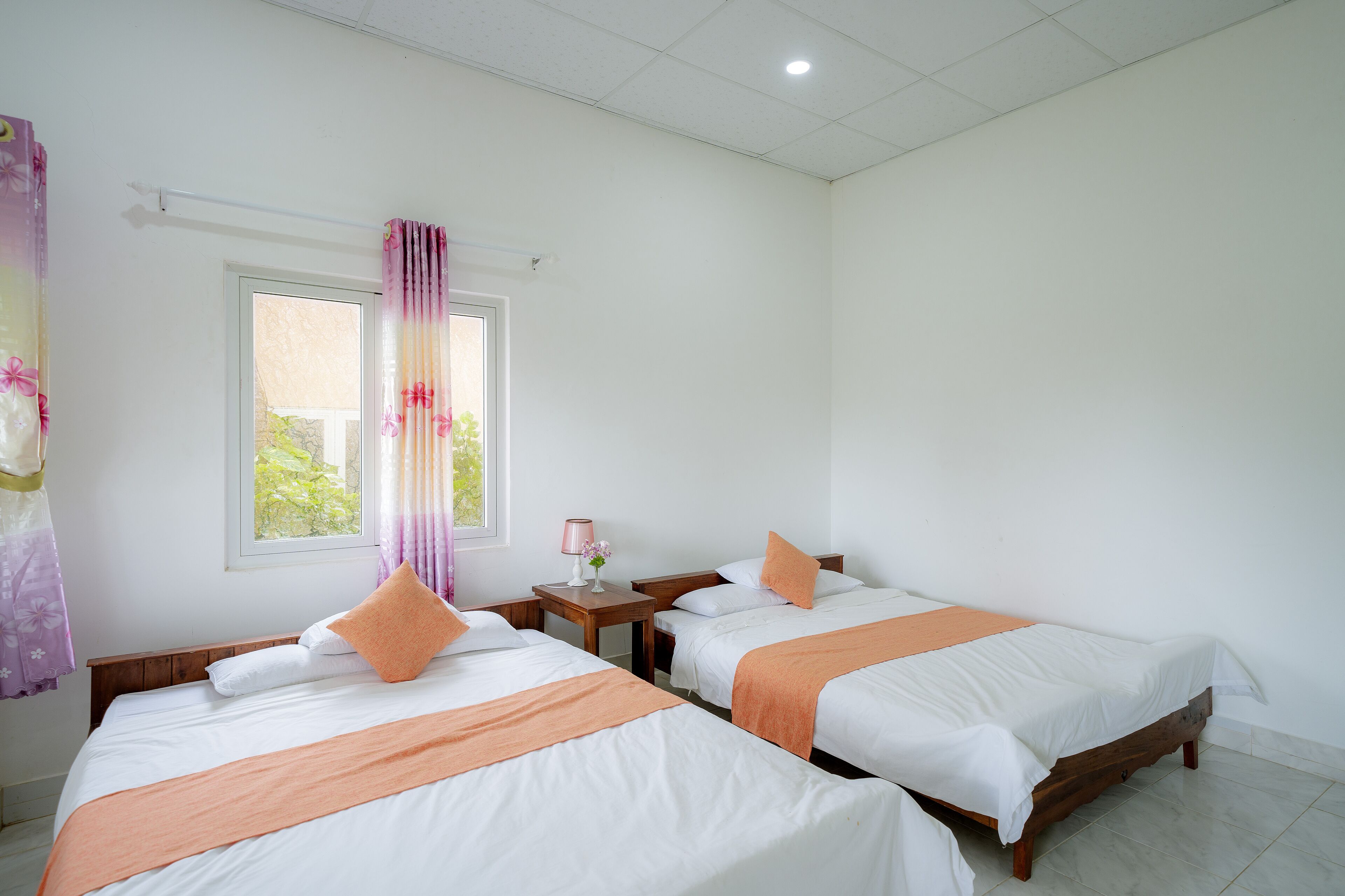 A hotel room with two single beds, a window with curtains, a bedside table with a lamp, and a view of greenery outside.