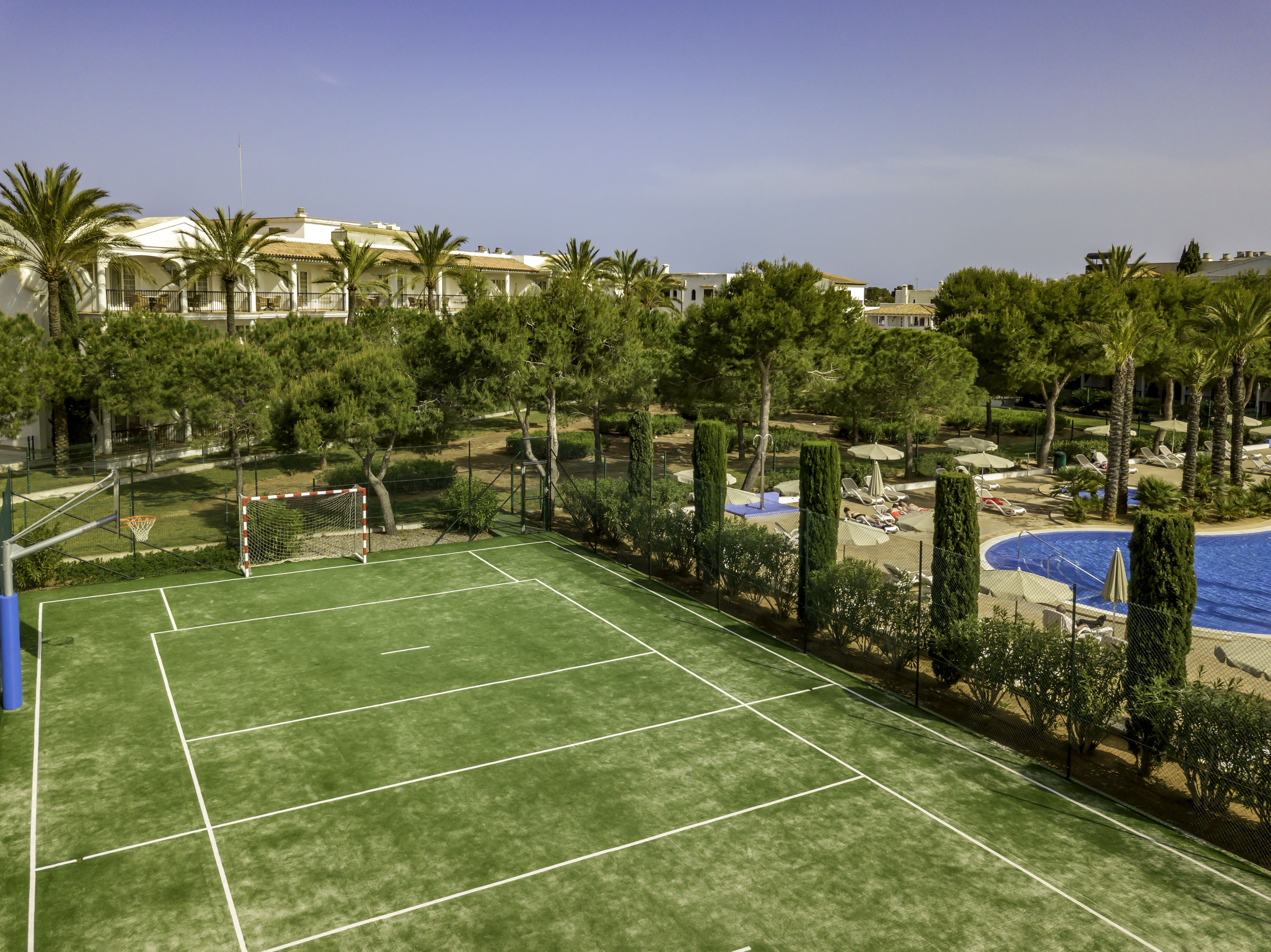 A tennis court with a net, surrounded by palm trees and a pool area with lounge chairs.