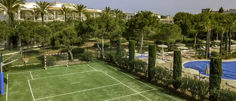 A tennis court with a net, surrounded by palm trees and a pool area with lounge chairs.
