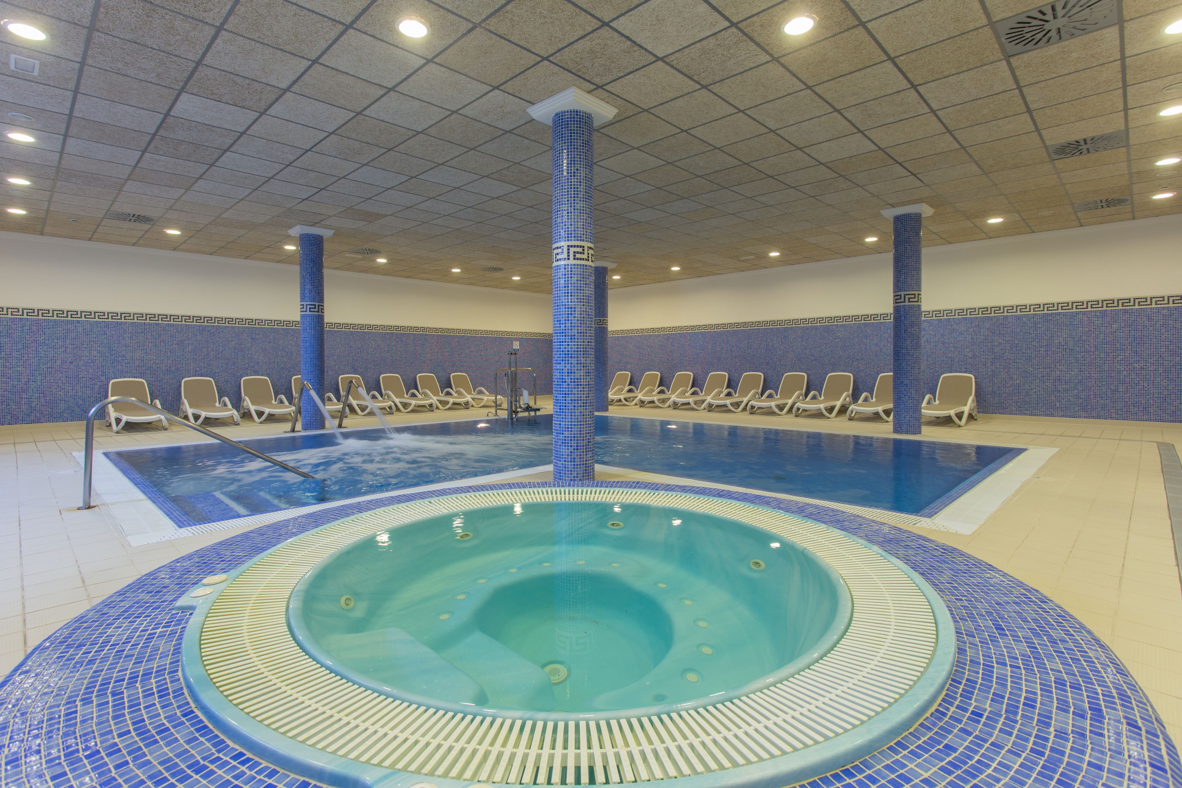 Una amplia piscina interior con un jacuzzi en el centro, rodeada de sillones de descanso.