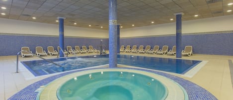 Una amplia piscina interior con un jacuzzi en el centro, rodeada de sillones de descanso.