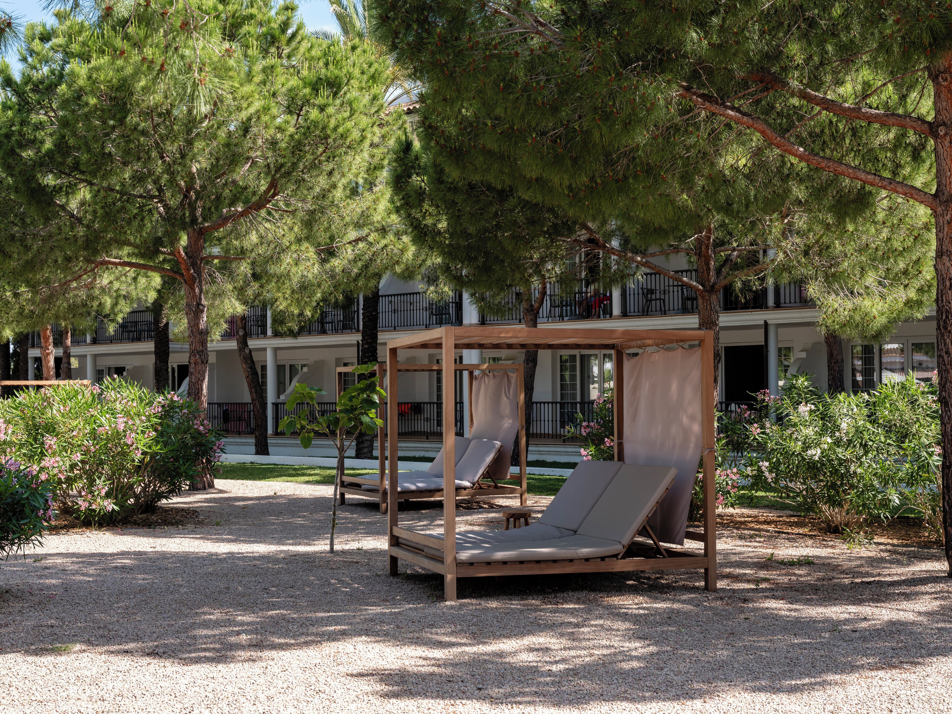 A serene outdoor seating area with wooden loungers and a pergola, surrounded by lush greenery and a building with balconies in the background.