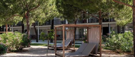A serene outdoor seating area with wooden loungers and a pergola, surrounded by lush greenery and a building with balconies in the background.